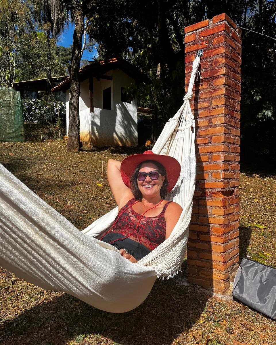 A Luciana, de chapéu vermelho, sorrindo deitada numa rede ao lado de uma pilastra de tijolos